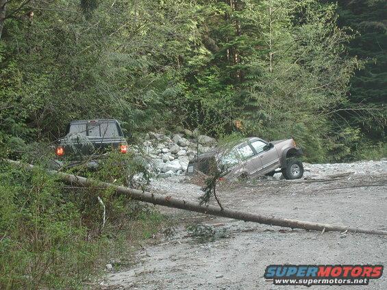 Stuck tan SUV and a pickup truck on a rocky path with a fallen tree.