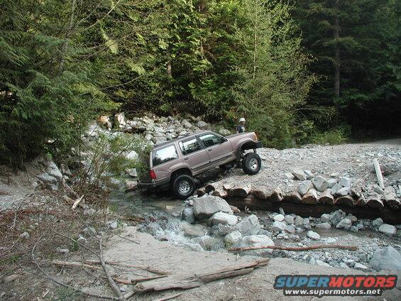 Lifted tan SUV driving through a creek over a log bridge in a forest.