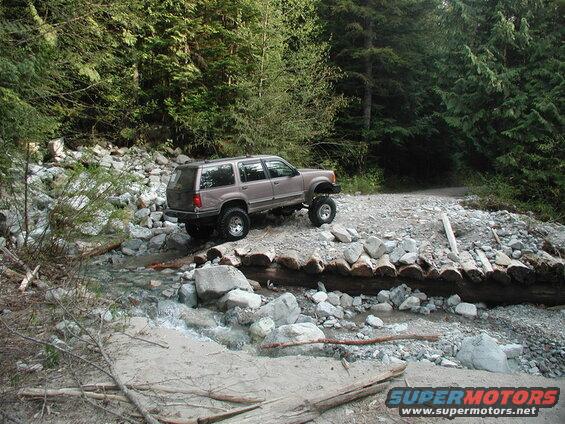 Lifted tan Ford Explorer crossing a stream on a log bridge in a forest.