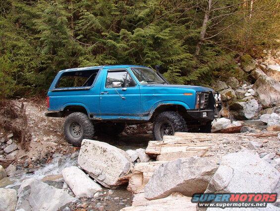 Blue 1980s Ford Bronco off-roading through a rocky stream.