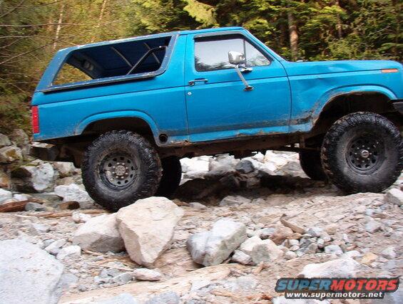 Side view of a blue 1985 Ford Bronco driving on a rocky trail.