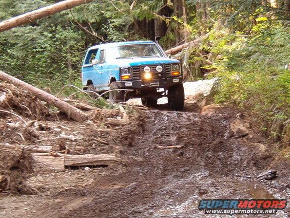 Blue 1985 Ford Bronco off-roading on a muddy, wooded trail.