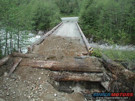 Damaged wooden bridge over a river with exposed logs and dirt.
