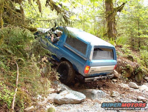 Blue Ford Bronco climbing a steep, rocky, wooded trail.