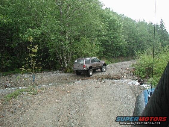 Lifted tan SUV crossing a shallow stream on a gravel road.