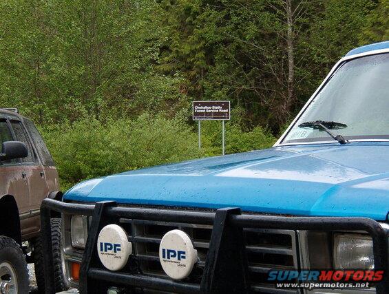 Blue 1985 Ford Bronco with bull bar and IPF lights, parked next to a brown SUV near a forest service road sign.