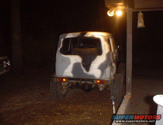 Rear view of a camo-painted Suzuki Samurai parked next to a pole with a light. Another vehicle is partially visible to the left