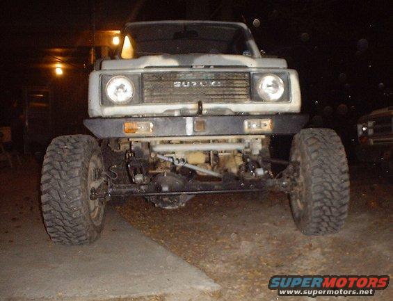 Suzuki Samurai with aftermarket bumper and large tires, front view. Another vehicle is parked to the right