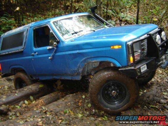 Blue 1985 Ford Bronco off-roading over logs and mud.