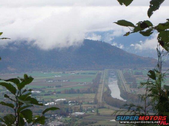 Valley with river, farmland, and mountain under cloudy sky, framed by foliage.