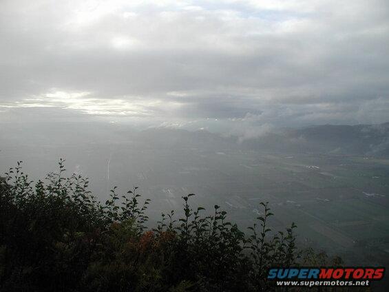 Overcast valley view with sun breaking through clouds and silhouetted foliage in foreground.