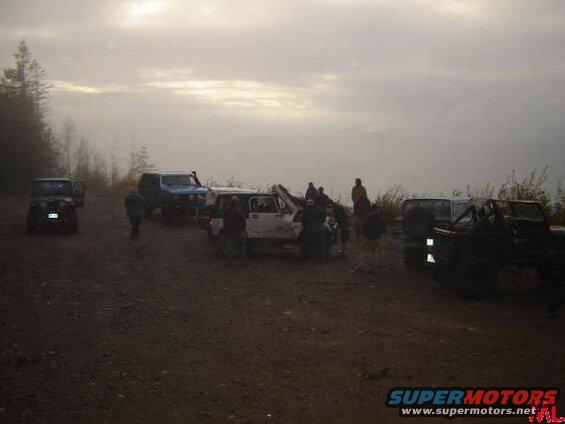 Off-road vehicles on a dirt road with people gathered around a white SUV.