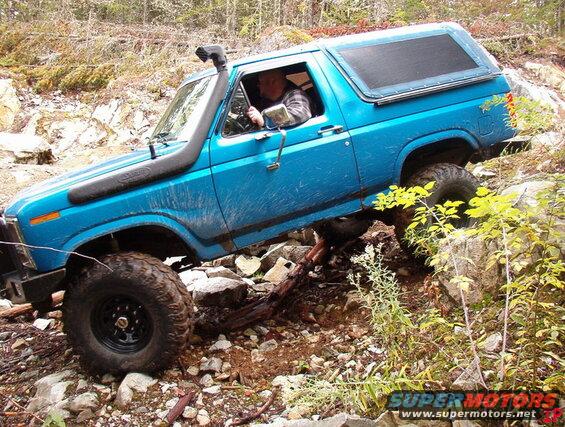 Blue 1985 Ford Bronco off-roading on a rocky trail with a snorkel.