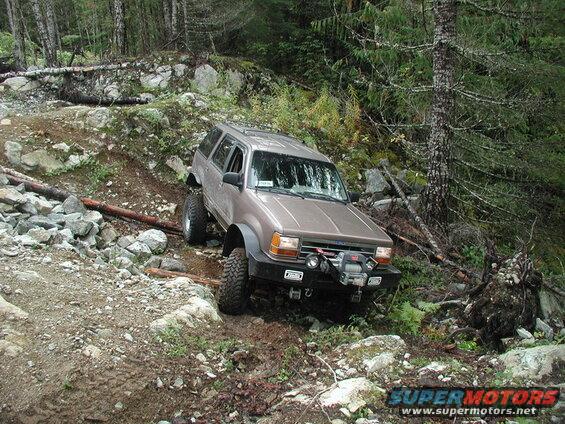 Tan SUV with off-road modifications on a rocky, wooded trail.