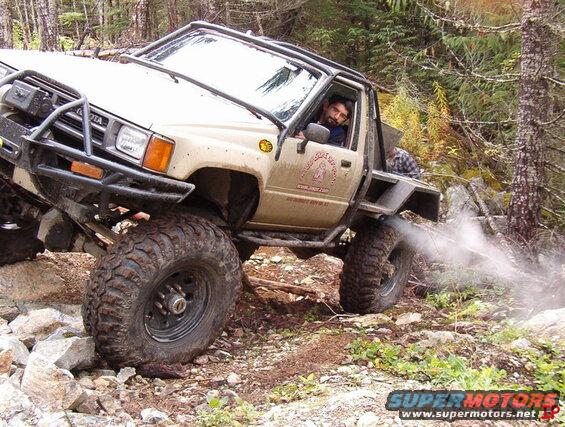 Tan Toyota pickup truck with large off-road tires on a rocky, wooded trail, driver visible.
