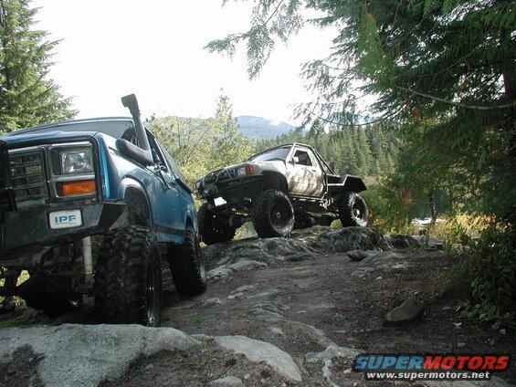 Two off-road trucks on a rocky trail with trees and mountains in the background.
