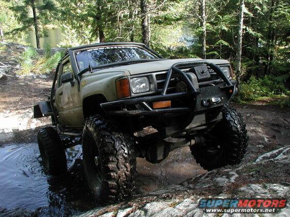 Lifted olive green Toyota 4x4 truck with large tires driving through water and rocks in a forest.