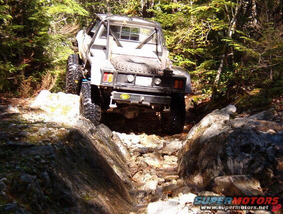 Off-road pickup truck with large tires and roll cage driving on a rocky forest trail.