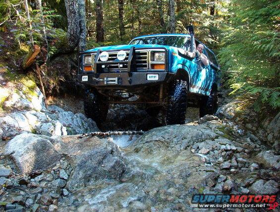 Blue Ford Bronco off-roading through a rocky stream in a forest.