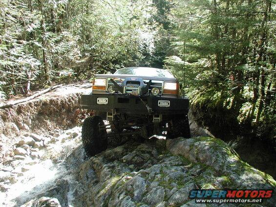 Modified dark brown SUV driving through a rocky stream in a forest.