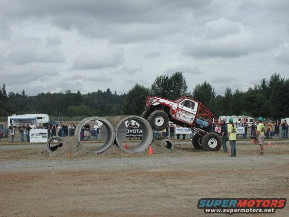 Monster truck with flames and red roll cage driving over concrete pipes.
