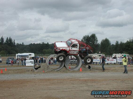 Custom off-road truck with red roll cage driving over concrete pipes with spectators in background.