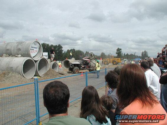 Custom off-road buggy airborne over dirt and concrete pipes at an event with spectators.