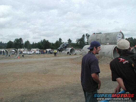 Dark SUV driving over large concrete pipes at an outdoor event.