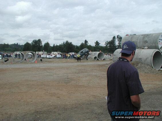 Dark SUV jumping over a concrete pipe obstacle at an outdoor event.