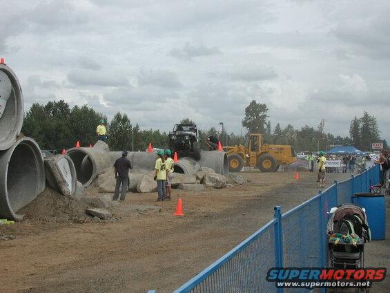 Black off-road vehicle on a pipe and rock obstacle course with a front-end loader and spectators.