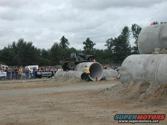 Modified pickup truck driving over concrete pipes with spectators watching.