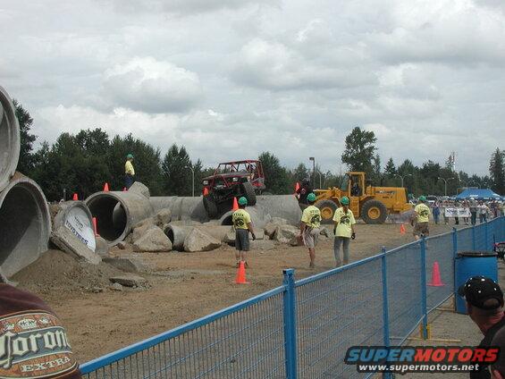 Red off-road buggy climbing over concrete pipes and rocks at an outdoor event.