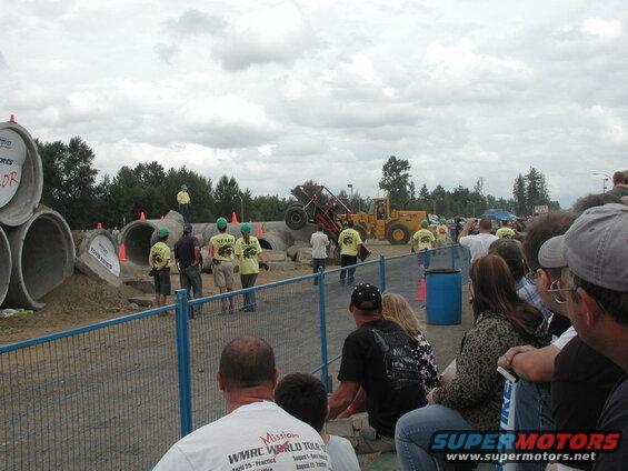 Custom off-road buggy airborne over concrete pipes at an event, with staff and spectators watching.