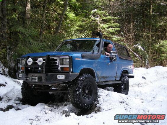 Blue Ford Bronco driving on a snowy forest trail with a driver visible.