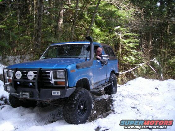 Blue 1985 Ford Bronco with snorkel and off-road bumper driving in snow.