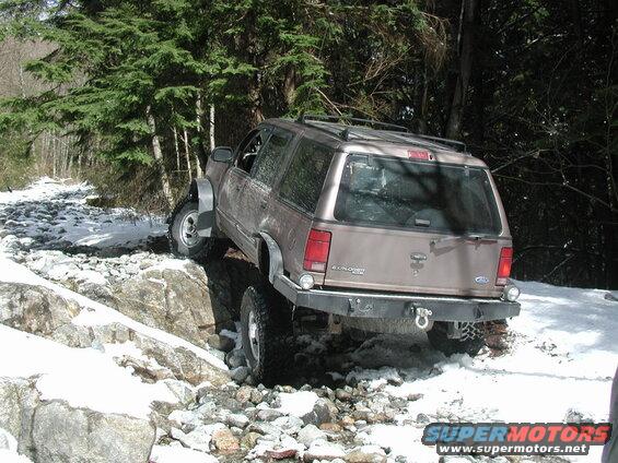 Tan Ford Explorer off-roading on a rocky, snowy path with trees.