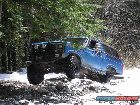 Lifted blue 1980s Ford Bronco off-roading on a rocky, snowy forest trail.