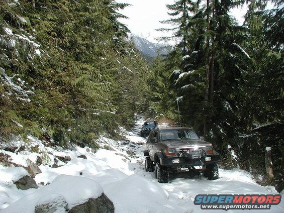 Brown SUV with off-road modifications driving on a snowy forest trail.
