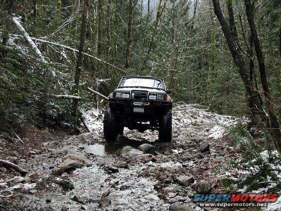 White 1980s SUV with off-road tires and lights on a snowy, rocky forest trail.
