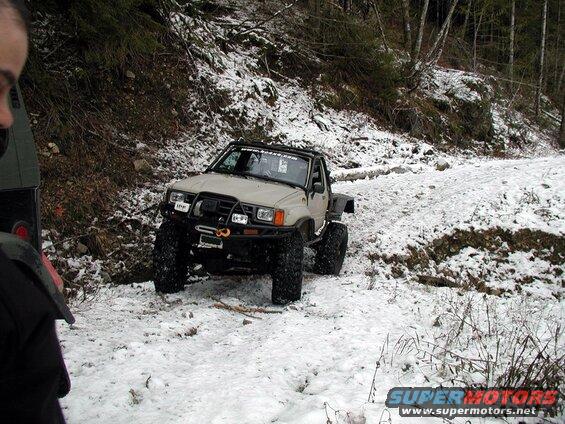 Tan off-road pickup truck with large tires on a snowy wooded trail.