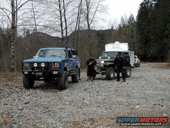 Blue Ford Bronco and black Jeep Wrangler with trailer parked on gravel road in woods.
