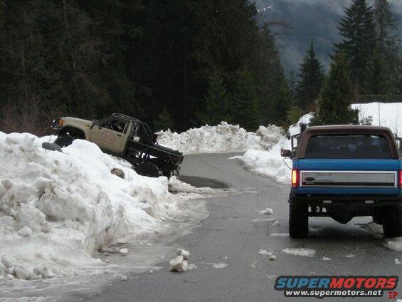 Blue Ford Bronco on a snowy road with a tan pickup truck stuck in snow.