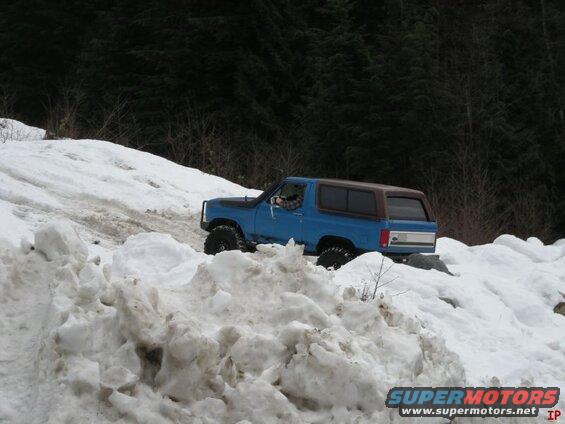 Blue first-generation Ford Bronco with brown top driving on a snowy road in a forest.