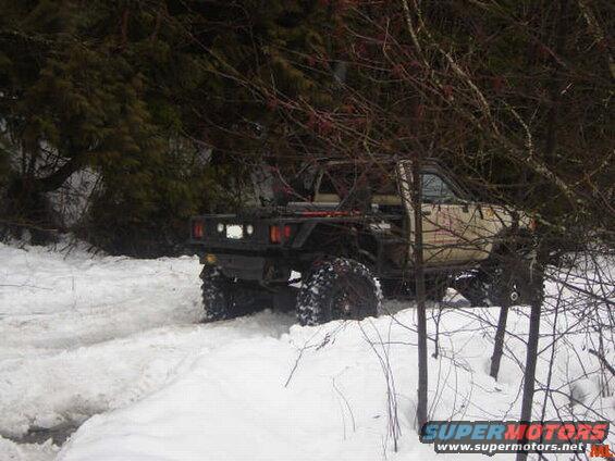 Modified pickup truck with large tires driving through snow in a forest.