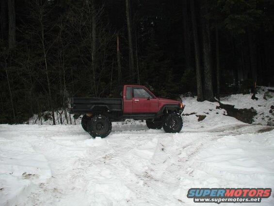 Red flatbed pickup truck with large off-road tires in a snowy forest.