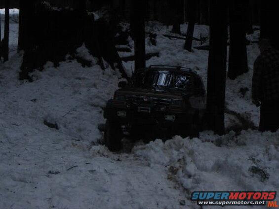 Dark SUV stuck in snow in a forest with a person standing nearby.