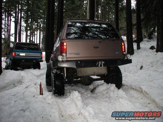 Two lifted SUVs driving on a snowy path in a forest.