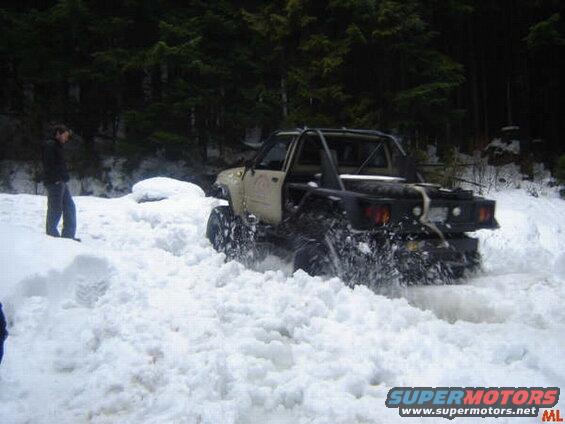Tan off-road truck with roll cage driving through deep snow in woods.