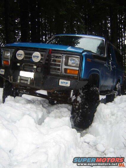 Blue Ford Bronco off-roading in deep snow with IPF lights and winch.