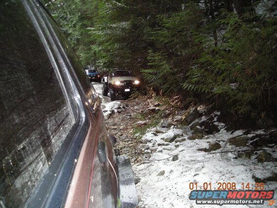 Off-road vehicles on a snowy forest trail, seen from the side of a dark SUV.